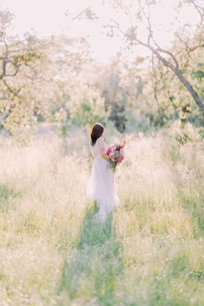 La hermosa foto lateral de la novia con el tatuaje de hombro negro en el maxi vestido blanco de boda sosteniendo el ramo y caminando en el bosque de primavera . — Foto de Stock