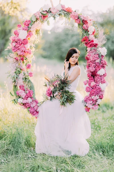 Porträttet av söt brud hålla buketten av blommor och sitter i bröllop pionerna arch i fältet soliga. — Stockfoto