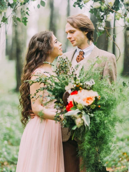 Närbild porträtt av de nygifta ser in i varandras ögon. Bruden med långt lockigt hår håller stor färgstark bukett. — Stockfoto