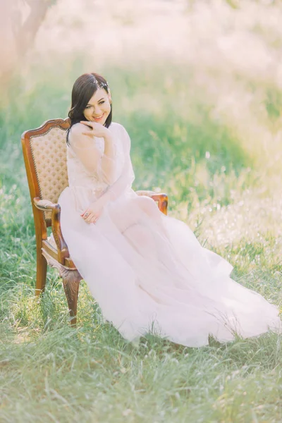 緑のスプリング フィールドの古いモダンな椅子に座って白いロング ・ ドレスで笑顔の女性のフルレングスの写真. — ストック写真