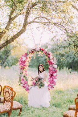 Güzel gelin buketi ile güneşli alan arka plan ağaç üzerinde asılı düğün çiçek Arch oturuyor.
