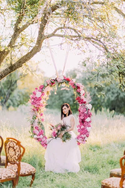 Την όμορφη νύφη με το μπουκέτο κάθεται στην αψίδα λουλούδι γάμο κρέμεται από το δέντρο στο βάθος του πεδίου ηλιόλουστος. — Φωτογραφία Αρχείου