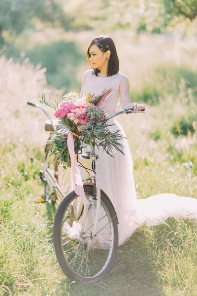 Η νύφη με το μακρύ λευκό φόρεμα και τα αξεσουάρ για τα μαλλιά κοιτάζοντας δεξιά πλευρά και κρατώντας το ποδήλατο με το μπουκέτο με πολύχρωμα λουλούδια. — Φωτογραφία Αρχείου