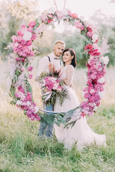 Newlyweds are holding the bouquet of peonies and standing behind the wedding peonies arch in the sunny spring field. Close-up vertical photo. — Stock Photo, Image