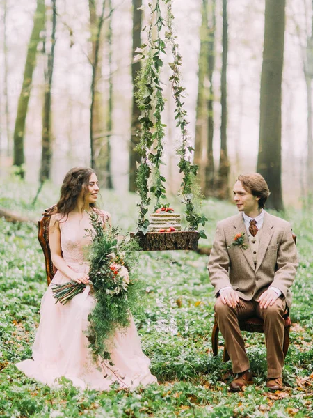 A linda foto dos recém-casados olhando uns para os outros enquanto sentados nas cadeiras entre o toco pendurado com a esponja de casamento bolo verde e branco sobre ele. A composição florestal . — Fotografia de Stock