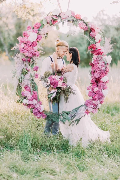The close-up portrait of the smiling newlyweds holding the bouquets of flowers and standing behind the wedding peonies arch in the sunny park. — Stock Photo, Image