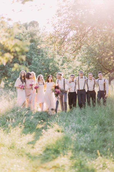 Foto dei migliori uomini vestiti moderno, sposa, damigelle d'onore che tengono il mazzo di fiori e lo sposo che trasporta la bicicletta con i fiori rosa. Composizione sul campo . — Foto Stock