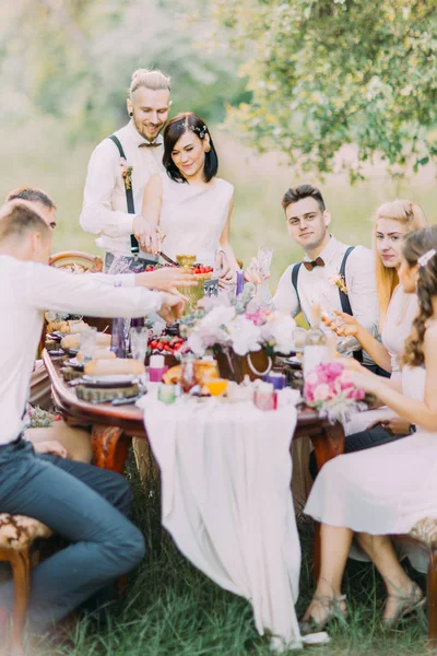 The lovely portrait of the newlyweds cutting their wedding cake and their guests. The table setting of the wedding dinner located in the sunny field.