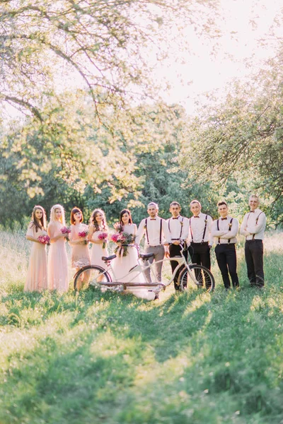 The group photo of the groom and his best men, the bride with the bridemaids behing the white bicycle at the background of the sunny green forest.