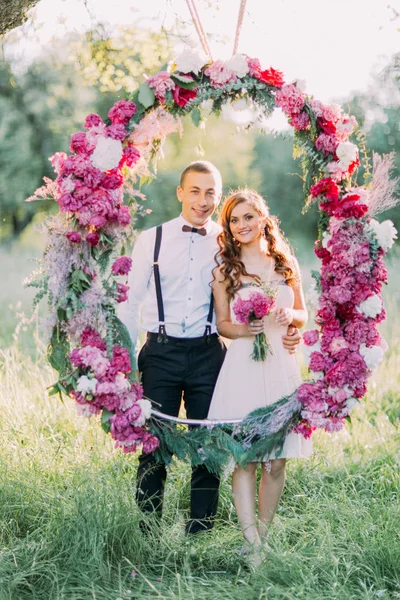 A dama de honra e o melhor homem estão posando através do arco de flores do casamento no campo ensolarado . — Fotografia de Stock