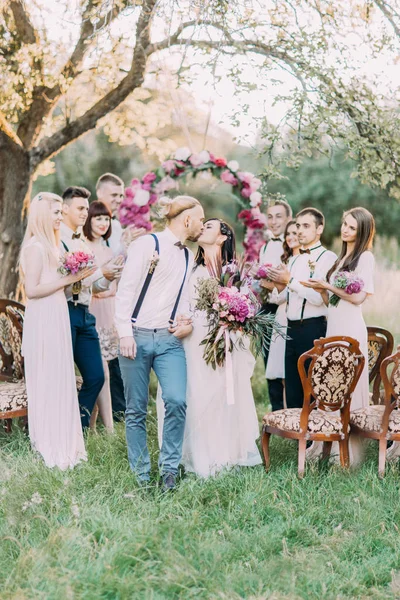 De verticale foto van de mooie ceremonie in het zonnige veld. De mooie pasgetrouwden zijn kussen op de achtergrond van hun gasten en de huwelijksboog pioenrozen. — Stockfoto