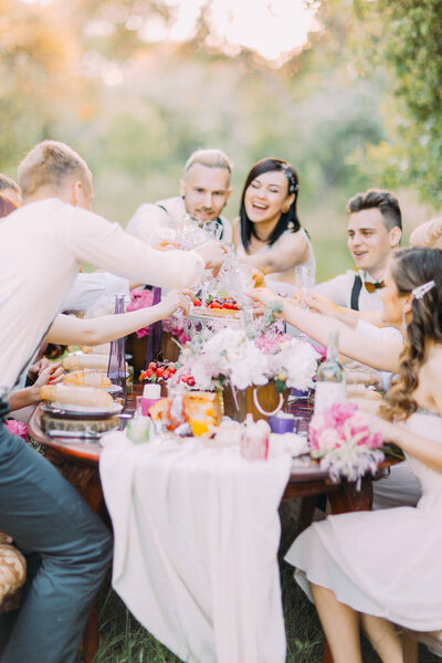 Happy close-up portrait of the delightful newlyweds and guests clinking their glasses and having fun at the setting table full of food and lovely flowers in the forest.