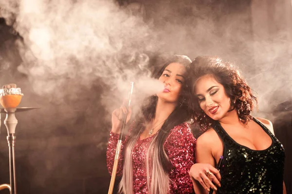 Dos mujeres jóvenes y guapas fumando narguile en un bar. Mujeres en vestidos de escote profundo y con maquillaje brillante . — Foto de Stock