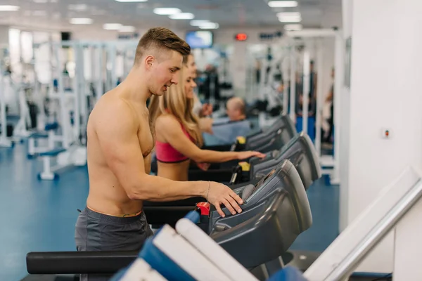 Ein junger Mann und zwei Frauen laufen auf Laufbändern im Fitnessstudio. Blondes Mädchen und Mann geben Lauftempo vor. — Stockfoto