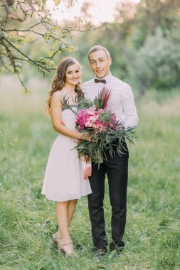 Büyük buket ve onunla orman arka plan sarılma gülümseyen en iyi adam tutarak diz boyu elbiseli gülümseyen nedime tam-yüksekliği portresi.