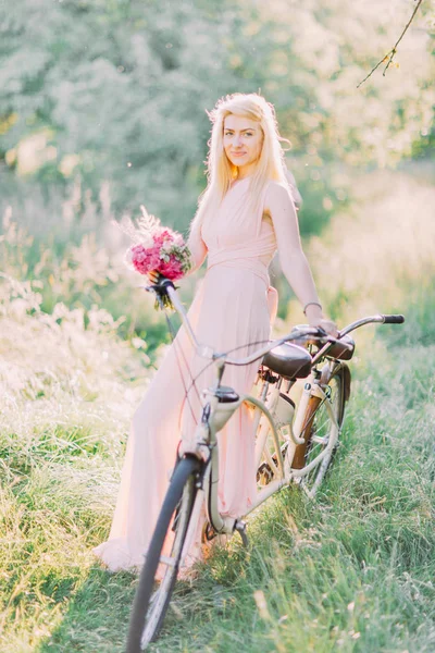 ピンクの花束を押しながら日当たりの良い森の中の自転車の上に座って長い光ピンクのドレスで花嫁介添人の垂直写真. — ストック写真