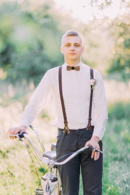 Siyah pantolon, beyaz gömlek, karanlık pantolon askısı ve ahşap kelebek en iyi adam Bisiklet güneşli ormanda tutuyor.