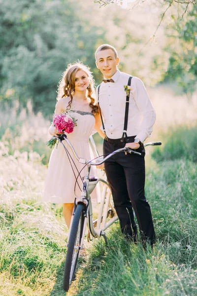 A foto vertical da dama de honra no vestido nu curto segurando o buquê das flores rosa e o melhor homem no terno moderno carregando a bicicleta na floresta ensolarada . — Fotografia de Stock