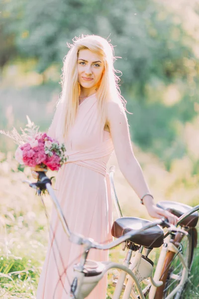 O lindo retrato da dama de honra loira no vestido rosa longo segurando o buquê e de pé perto da bicicleta no campo ensolarado . — Fotografia de Stock