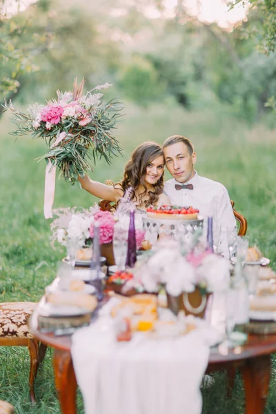 The bridesmaid is holding up the lovely bouquet, hugging with the best man and they are sitting at the wedding table, having dinner in the sunny field. — Stock Photo, Image