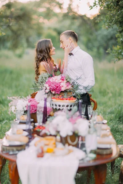 O lindo retrato lateral da dama de honra acariciando o rosto do melhor homem e de pé atrás da mesa de casamento borrada no campo verde . — Fotografia de Stock