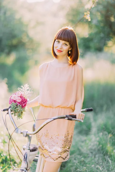 O retrato de close-up da dama de honra de corte de cabelo curta sorridente segurando o buquê rosa e carregando a bicicleta na floresta verde. O vestido é decorado por brilhos . — Fotografia de Stock
