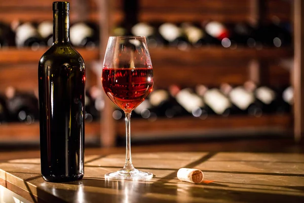 Tiro CLose-up de garrafa de vinho e vidro colocado em uma mesa de madeira. Prateleiras com garrafas de vinho no fundo . — Fotografia de Stock