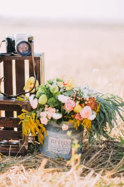 El cubo lleno de flores de colores se coloca cerca de las cajas de madera oscura con la cámara en el fondo del campo soleado . — Foto de Stock