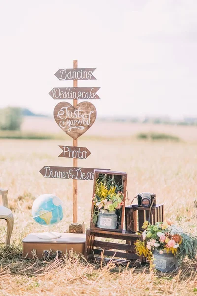 The vertical photo of the wooden plaques with the signs Wedding, photo, dancing placed near the wooden boxes with flowers, old camera and globe in the sunny field. — Stock Photo, Image