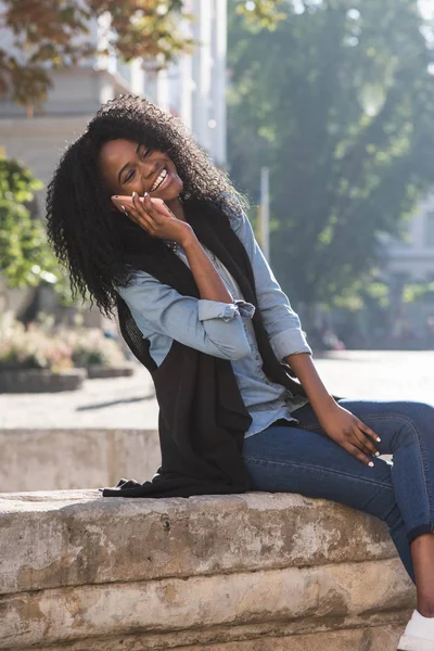 Encantadora chica afroamericana se ríe mientras habla a través del teléfono móvil. Ella está sentada en la fuente en el centro de la ciudad . — Foto de Stock