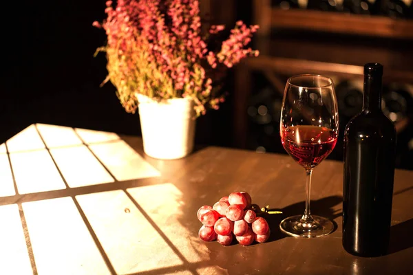 La composizione della bottiglia, del bicchiere di vino e del grappolo d'uva posto vicino al vaso di fiori sul tavolo nella stanza buia . — Foto Stock