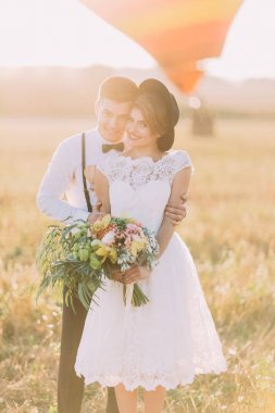 Gülümseyen damat büyük buket vintage giyinmiş gelinle güneşli alan arka geri sarılıyor.