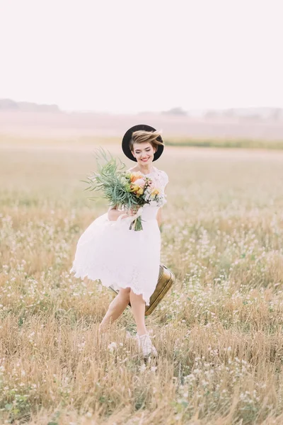 A noiva vestida vintage está andando no campo de primavera. Ela está segurando o buquê colorido e carregando a mala retro . — Fotografia de Stock