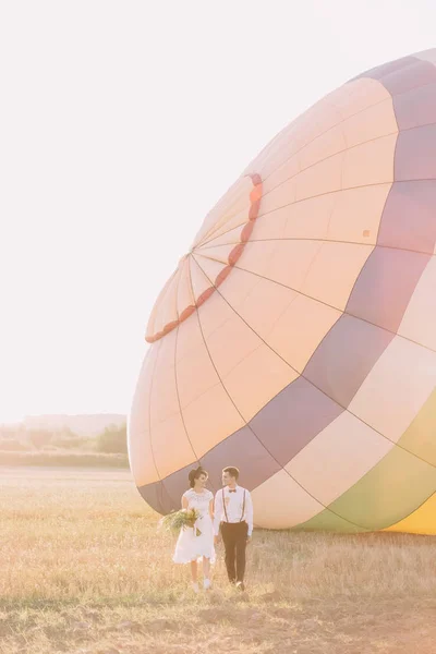 Szczęśliwi Nowożeńcy, trzymając się za ręce są spacery w polu słoneczny na tle kolorowe airballoon. — Zdjęcie stockowe