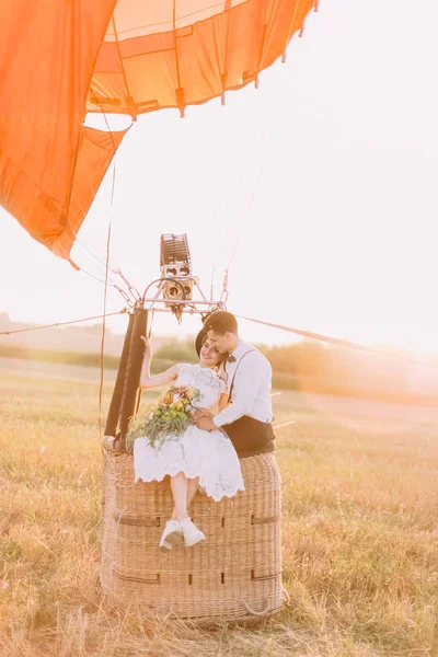 Вінтажні одягнені усміхнені молодята сидять у кошику на повітряній кулі, а наречена з букетом спирається на нареченого. Композиція поля — стокове фото