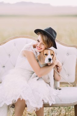 The smiling bride looking somewhere else is hugging the little dog sitting on the white vintage sofa at the background of the field. Close-up portrait. clipart