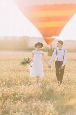 Gülümseyen vintage giyinmiş yeni evliler el ele tutuşarak ve güneşli alanına yürüyor. Gelin düğün buket airballoon geçmiş vasıl tutuyor..