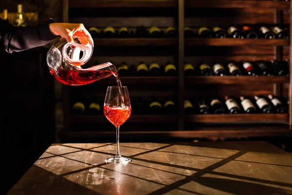 Red wine pouring into a glass from a carafe. Shelves with wine bottles on a background. — Stock Photo, Image