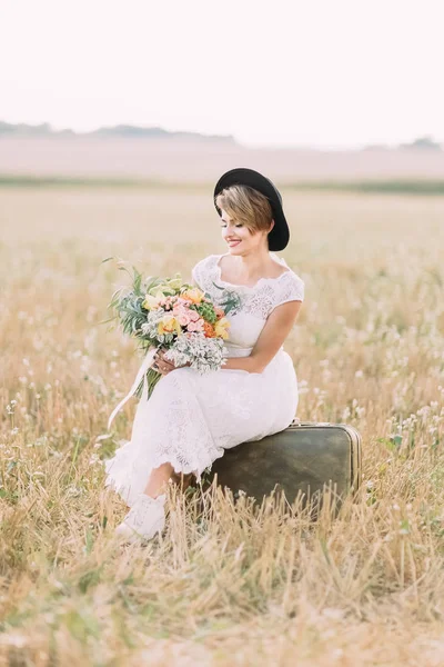 Vyn sida av härliga bruden sitter på vintage resväskan och tittar på den färgsprakande buketten med blommor i fältet soliga. — Stockfoto