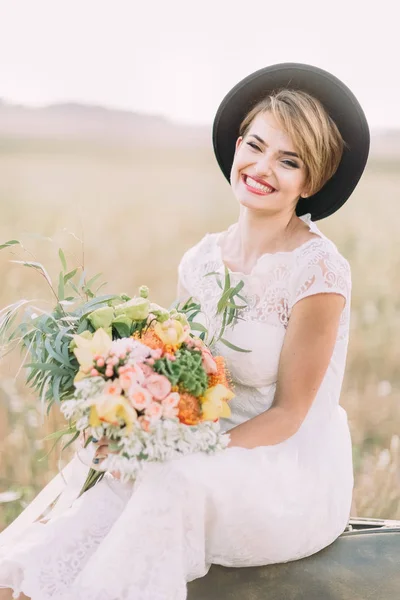 La sposa sorridente con il mazzo colorato grande che si siede sulla valigia d'annata tra il campo soleggiato . — Foto Stock