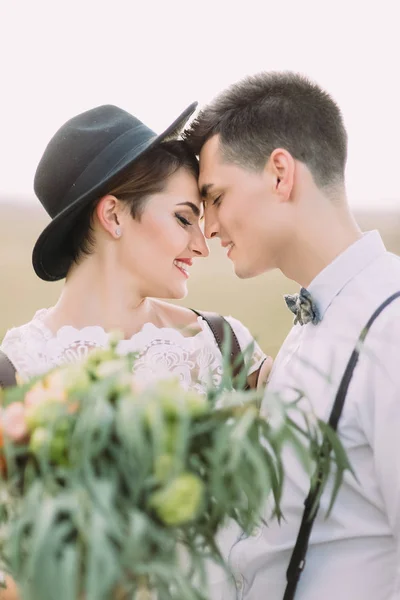 The close-up side portrait of the happy newlyweds standing head-to-head to each other behind the blurred bouquet of flowers. — Stock Photo, Image