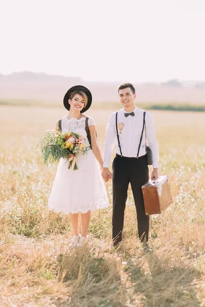 El retrato frontal de cuerpo entero de la pareja de recién casados vestidos vintage tomados de la mano y sonriendo a la cámara al fondo del campo soleado . — Foto de Stock