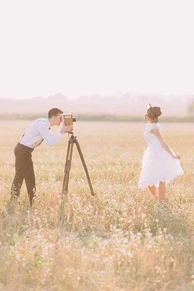 Het portret van de kant van de vintage gekleed pasgetrouwden in het veld. De bruidegom is het nemen van foto's terwijl de bruid is poseren. — Stockfoto