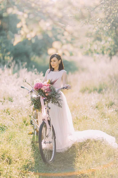 Κάθετη φωτογραφία της νύφης στο μακρύ άσπρο φόρεμα κρατώντας την ανθοδέσμη παιώνιες μεταφέρουν το ποδήλατο στο ηλιόλουστο πράσινο πεδίο. — Φωτογραφία Αρχείου