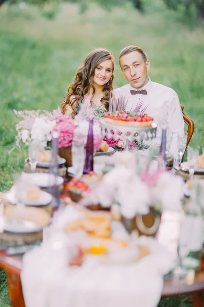 Il bel ritratto della giovane damigella d'onore e il testimone con la farfalla di legno sul collo seduto al tavolo delle nozze situato nella foresta soleggiata . — Foto Stock