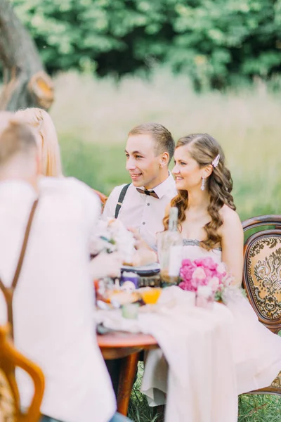 A foto vertical lateral da dama de honra no vestido rosa claro longo e o melhor homem com a borboleta de madeira sentada na mesa de casamento . — Fotografia de Stock