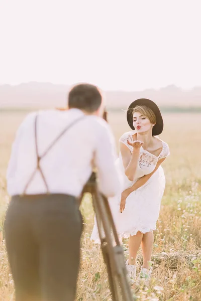 A visão traseira borrada do noivo tirando fotos da noiva enviando beijos no campo ensolarado . — Fotografia de Stock