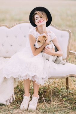 Gülümseyen vintage giyimli gelin siyah şapkalı güzel köpek sarılma ve güneşli alanındaki kanepede oturan.