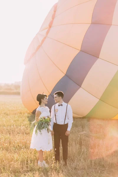 笑顔の花嫁が日当たりの良いフィールドである airballoon の backgrounhd に花束を保持する間に、手を繋いでいる新婚夫婦の素敵なフルレングス サイドの肖像画. — ストック写真