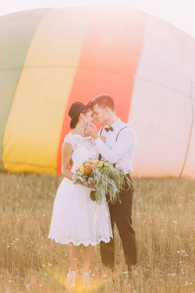 Os recém-casados felizes estão frente a frente na frente do balão de ar. A noiva está segurando o buquê de casamento enquanto o noivo está acariciando sua bochecha . — Fotografia de Stock